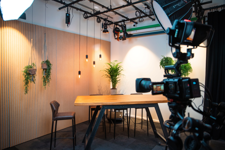 A modern studio setup with a wooden table, chairs, plants, warm lighting, and a camera on a tripod, ready for filming or broadcasting.