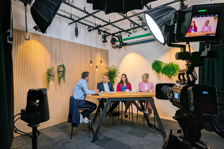 Four people sit at a wooden table in a studio with professional cameras and lighting. Green plants hang on the walls.
