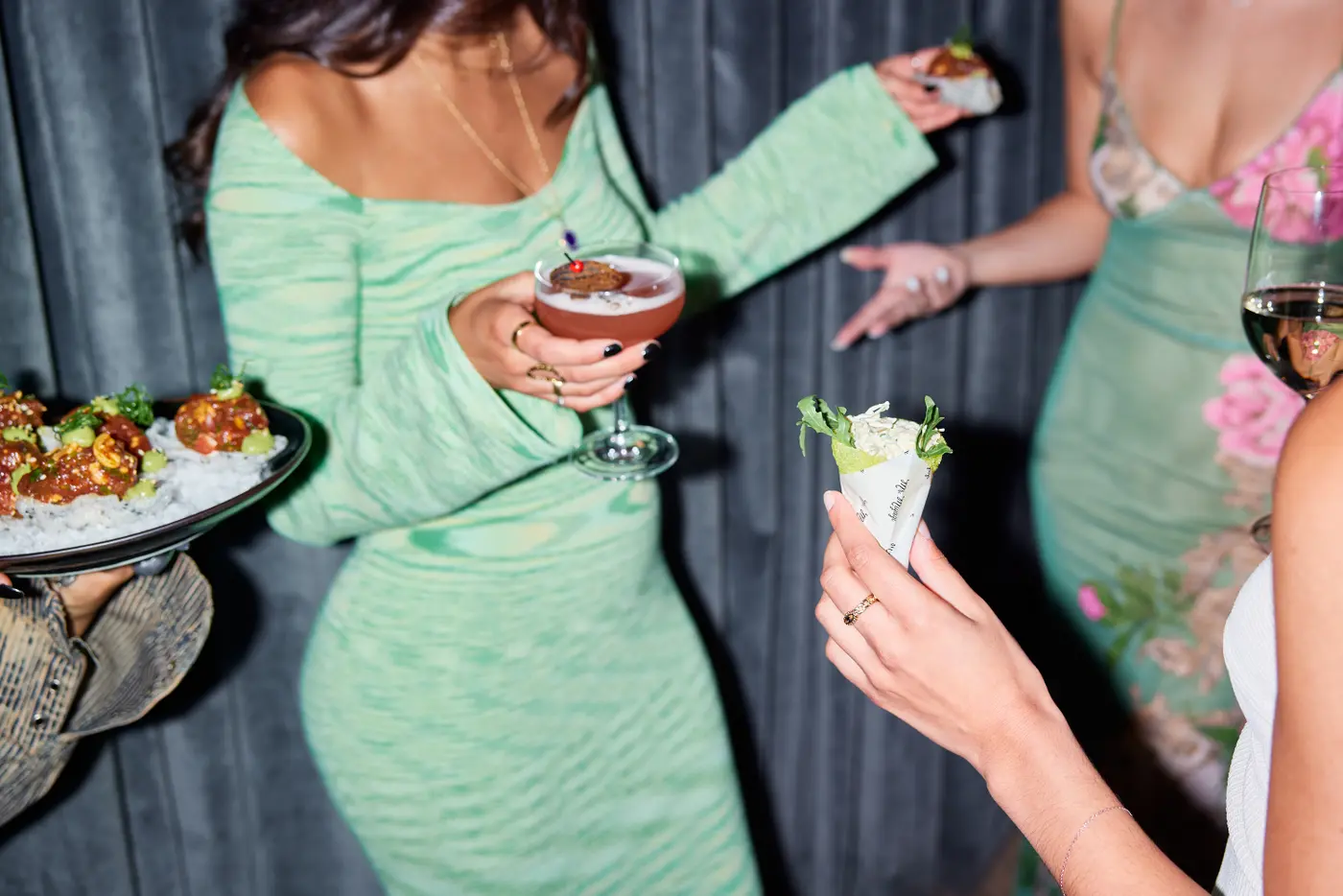 Party guests holding colourful cocktails and small bites against dark curtains