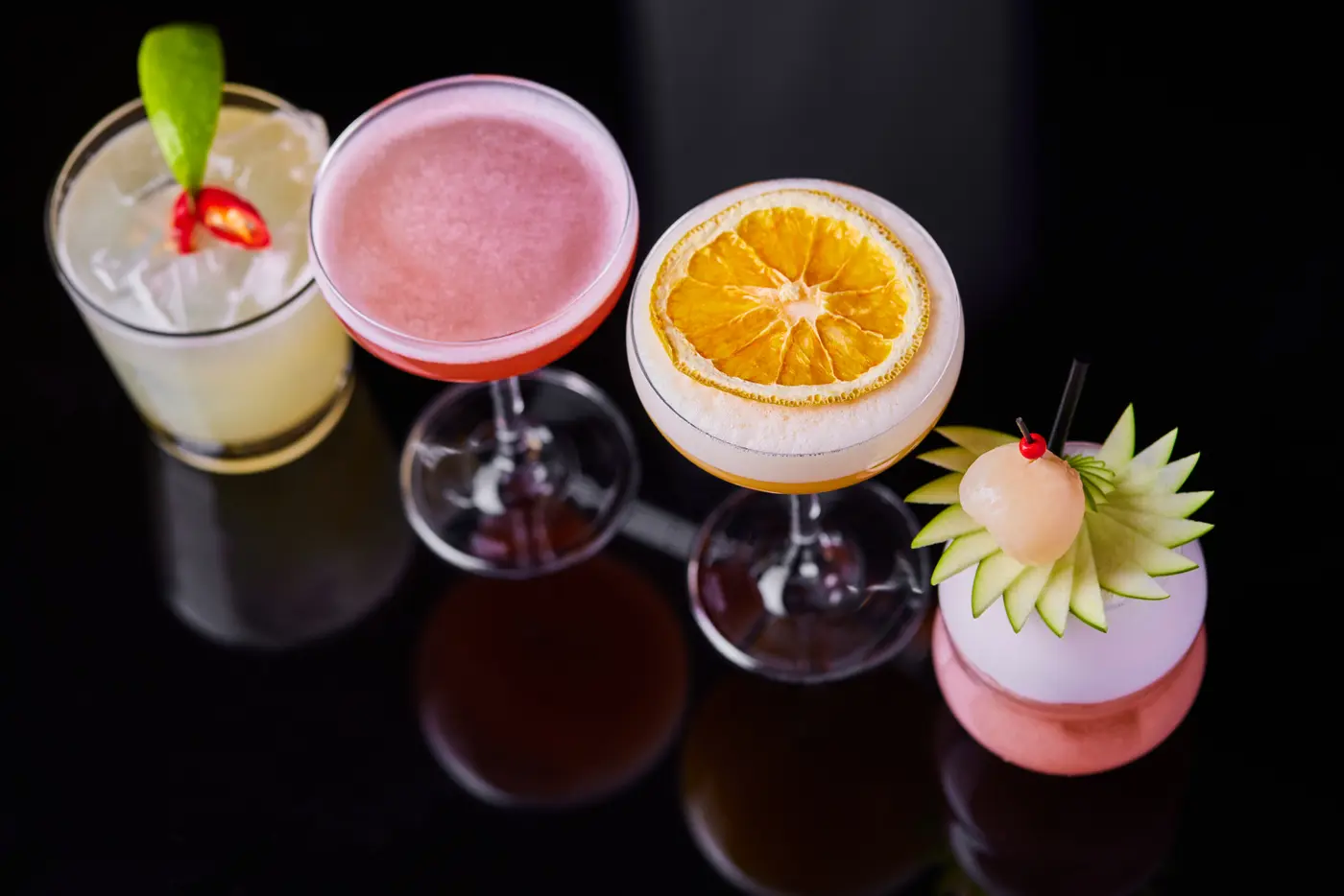 Four cocktails with fruit garnishes reflected on a glossy black surface