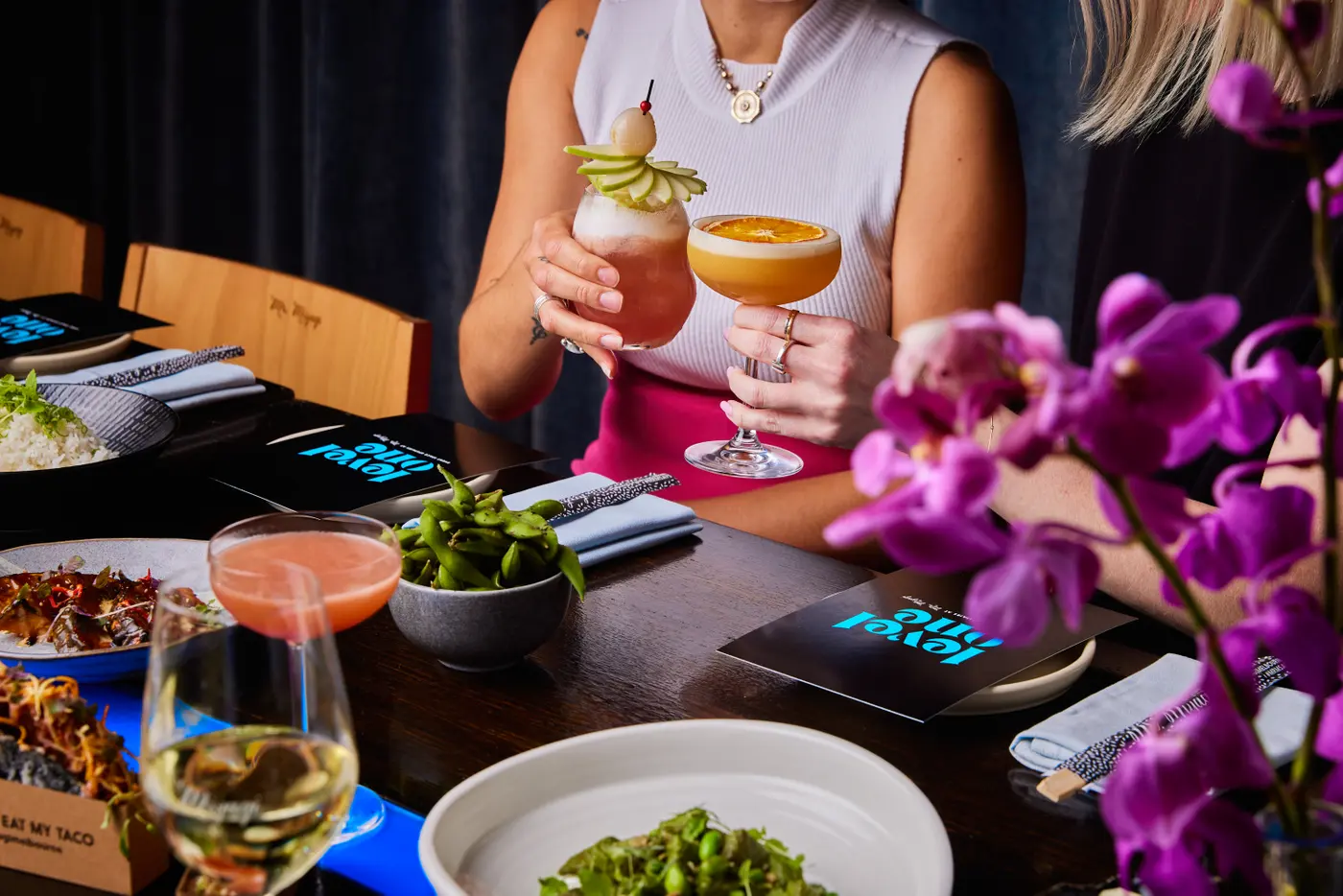 A guest holding two cocktails at a table with food and purple orchids