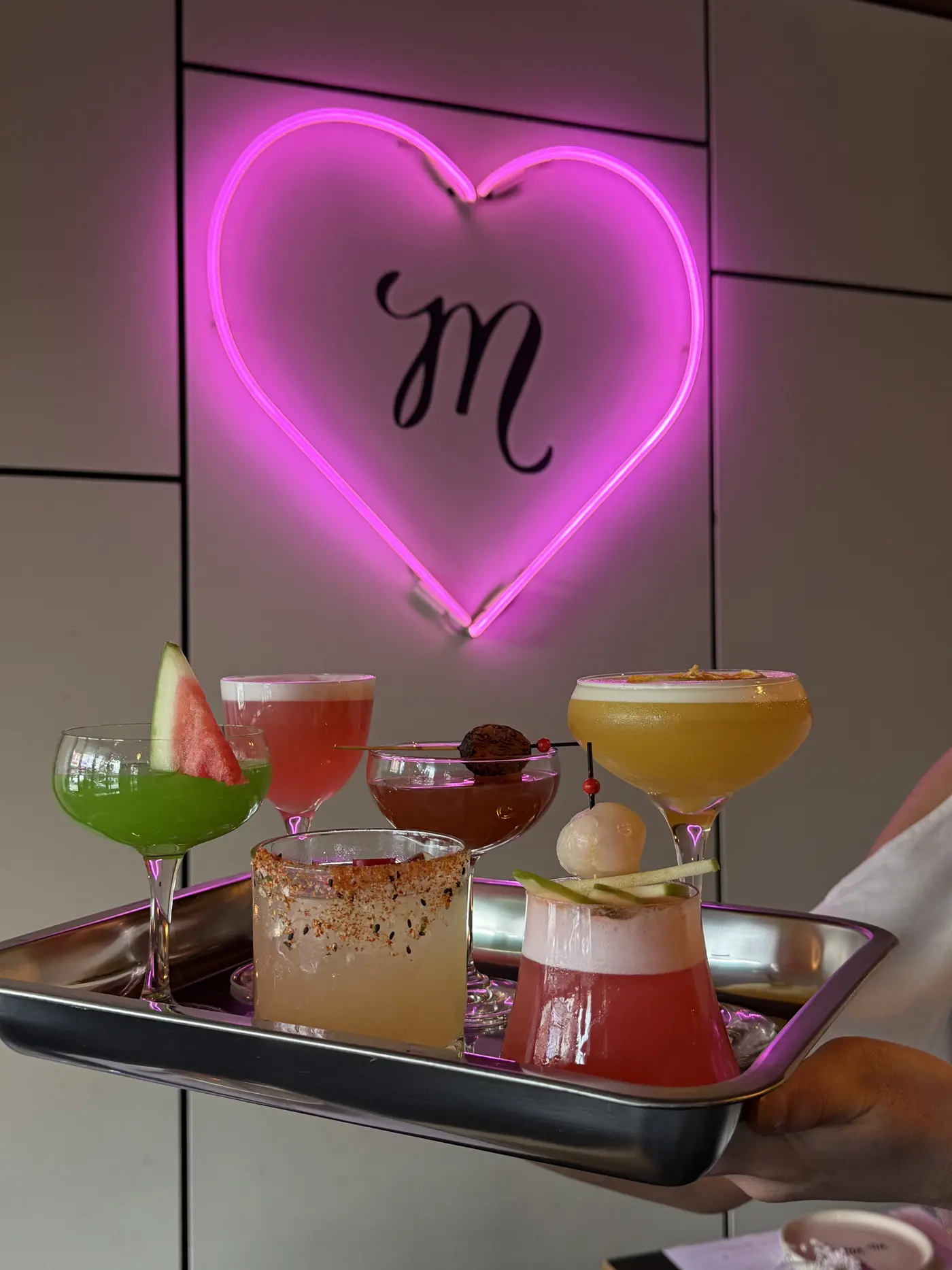 A tray of six colourful cocktails beneath a pink neon heart