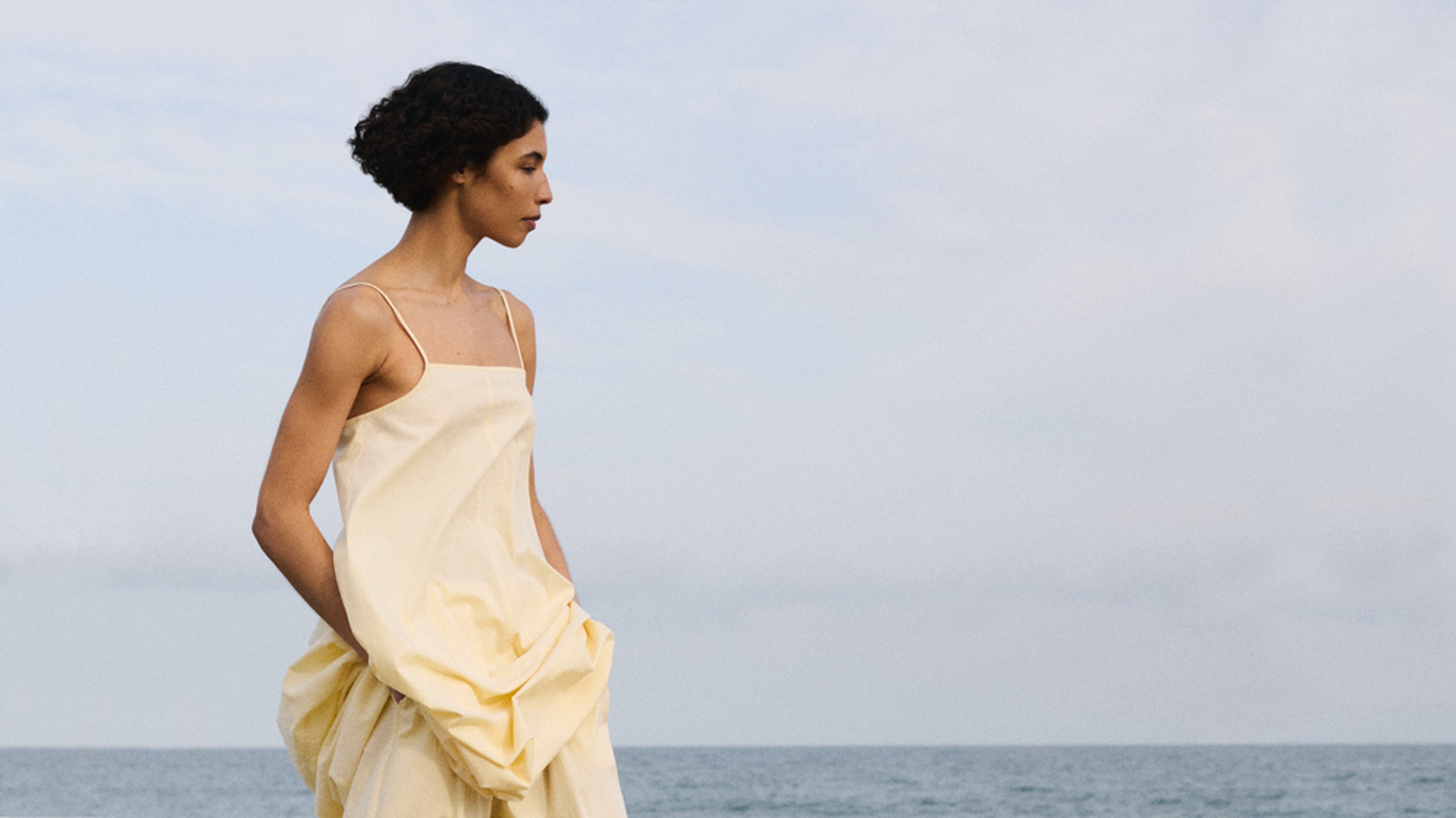 Woman in a flowing yellow dress stands by the sea, gazing into the distance under a cloudy sky.