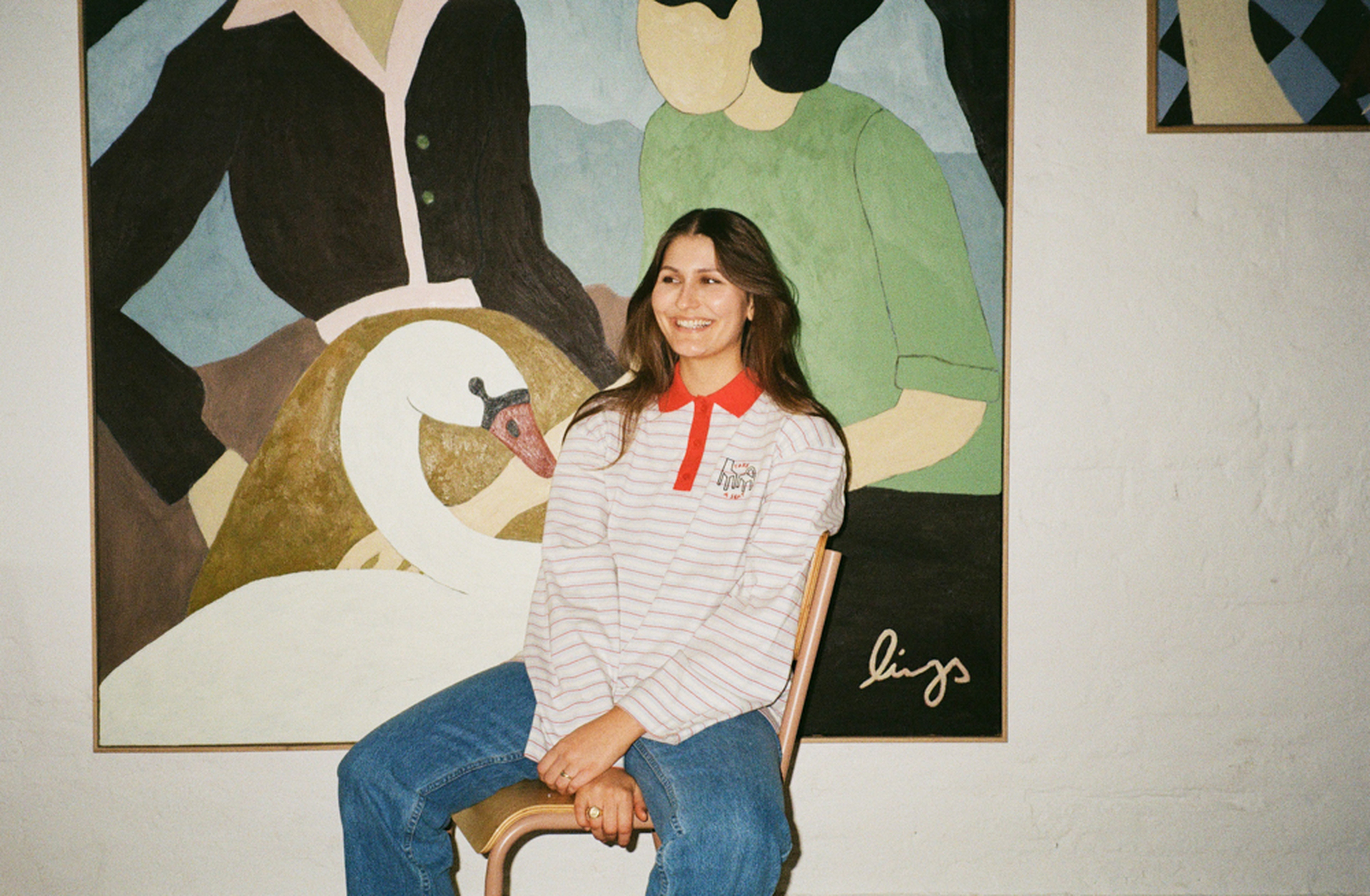 A woman sitting on a chair, smiling, with a colorful abstract painting of people and a swan behind her on the wall.