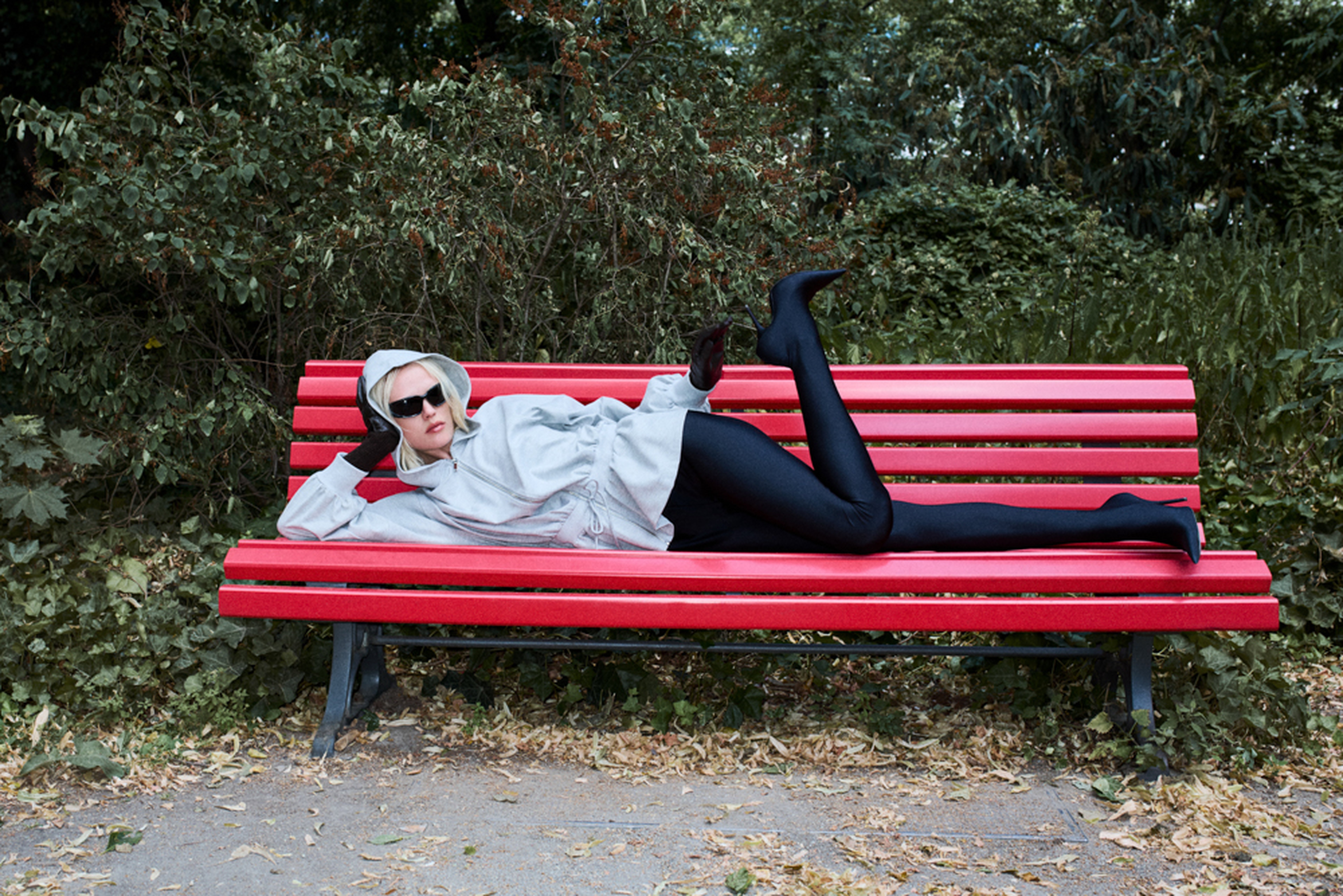Person in a gray hoodie, black tights, and sunglasses, lounging on a red park bench surrounded by greenery.