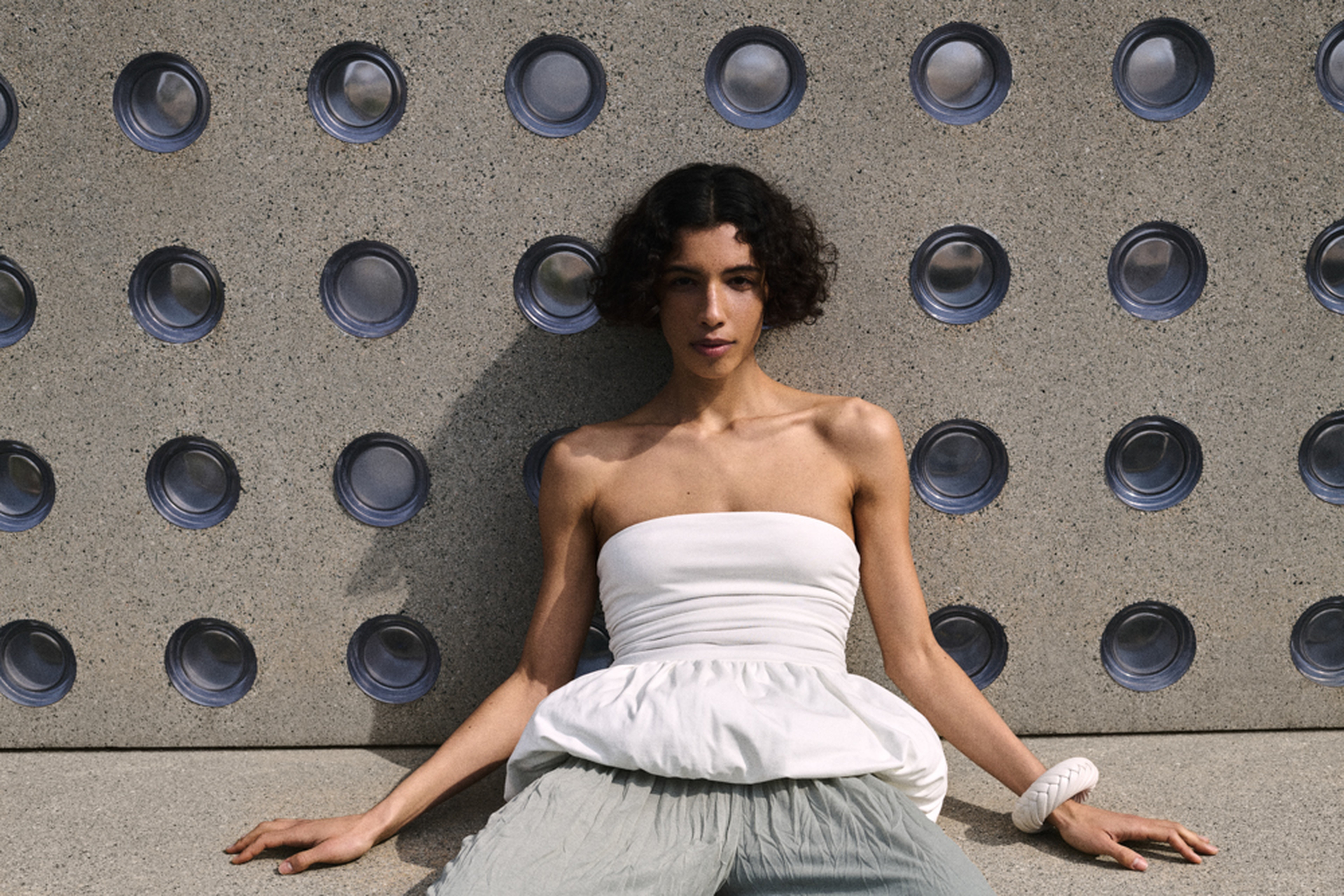 Woman in a strapless white top and light trousers leans against a textured wall with circular patterns, sitting on the ground.