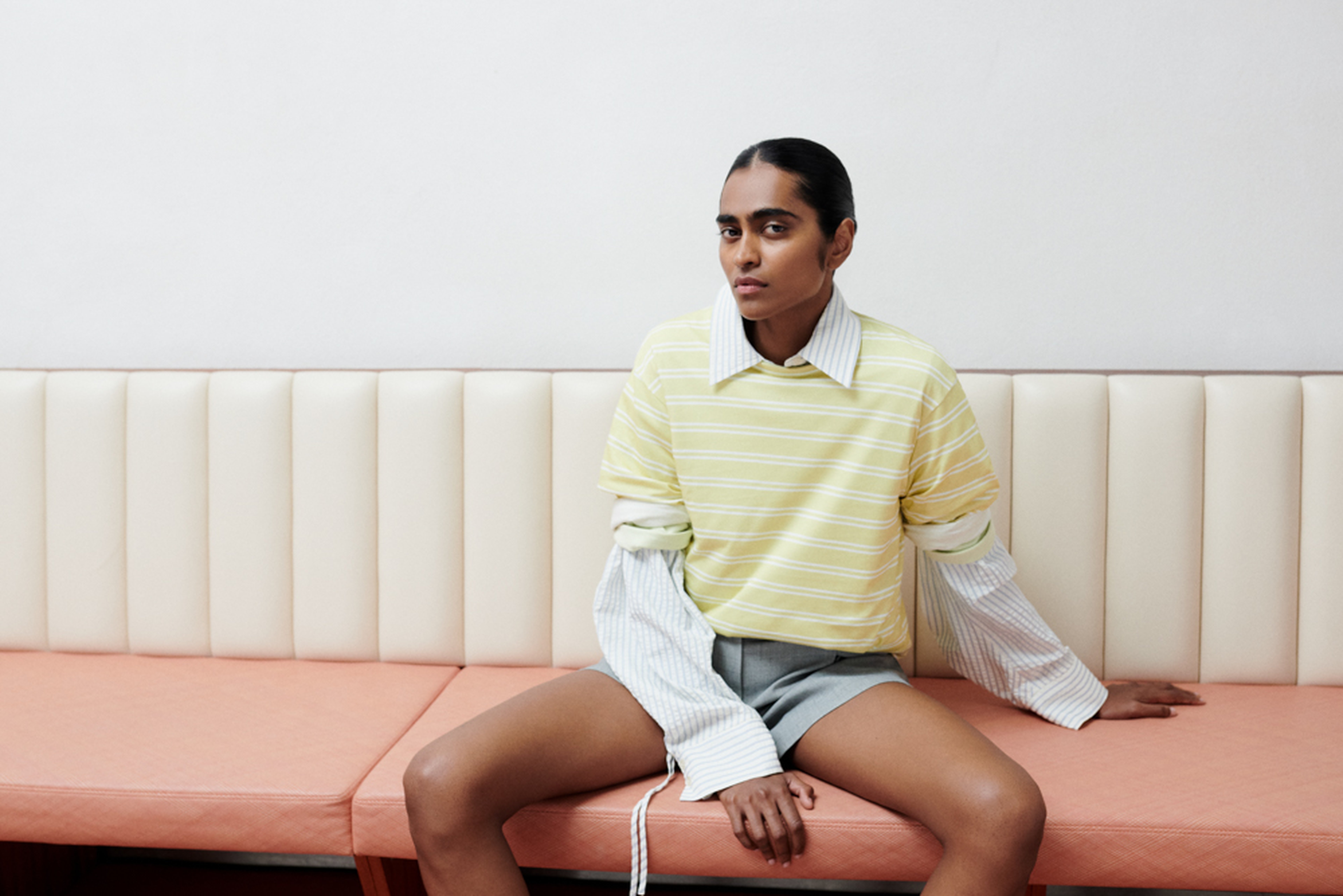 A person sitting on a bench wearing a yellow striped shirt over a white collared shirt, with gray shorts, against a white wall.