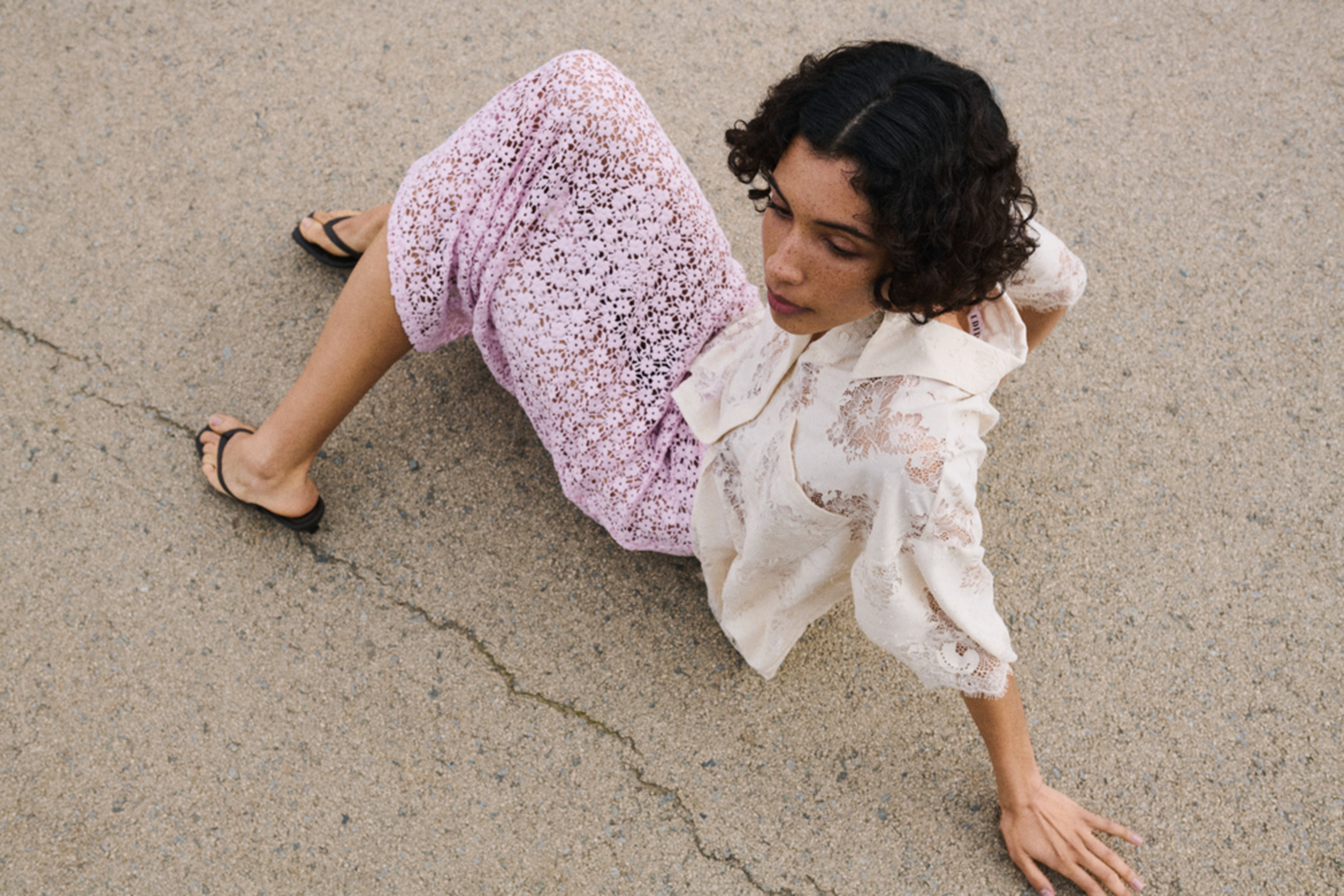 A woman in a lace blouse and pink skirt sits on a cracked pavement, looking upward.