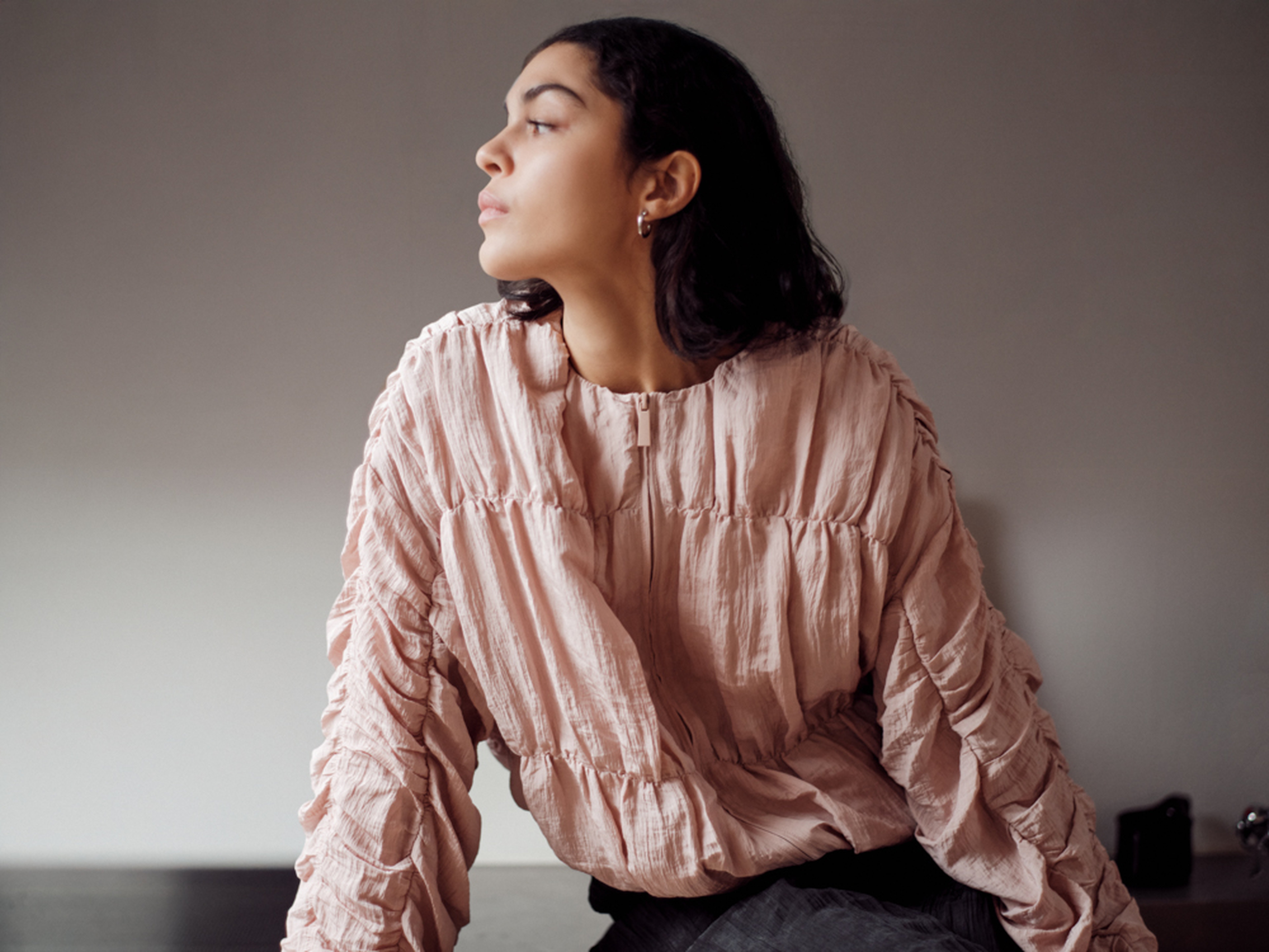 A person wearing a textured, pink blouse sits indoors, looking to the side with a contemplative expression, set against a plain background.