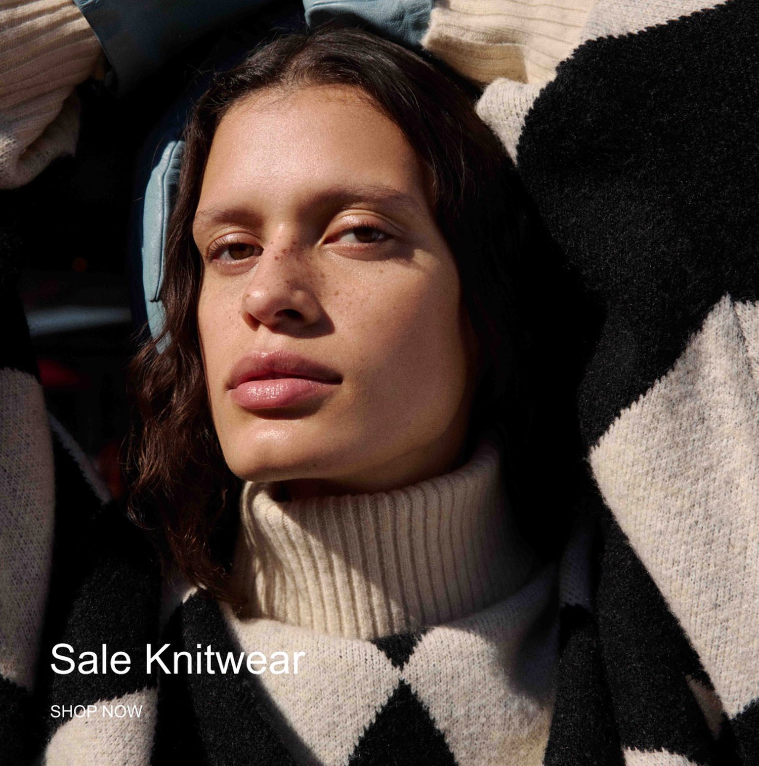 Person wearing a black and white patterned knit sweater with a high collar, looking at the camera. Text on image: "Sale Knitwear - SHOP NOW."