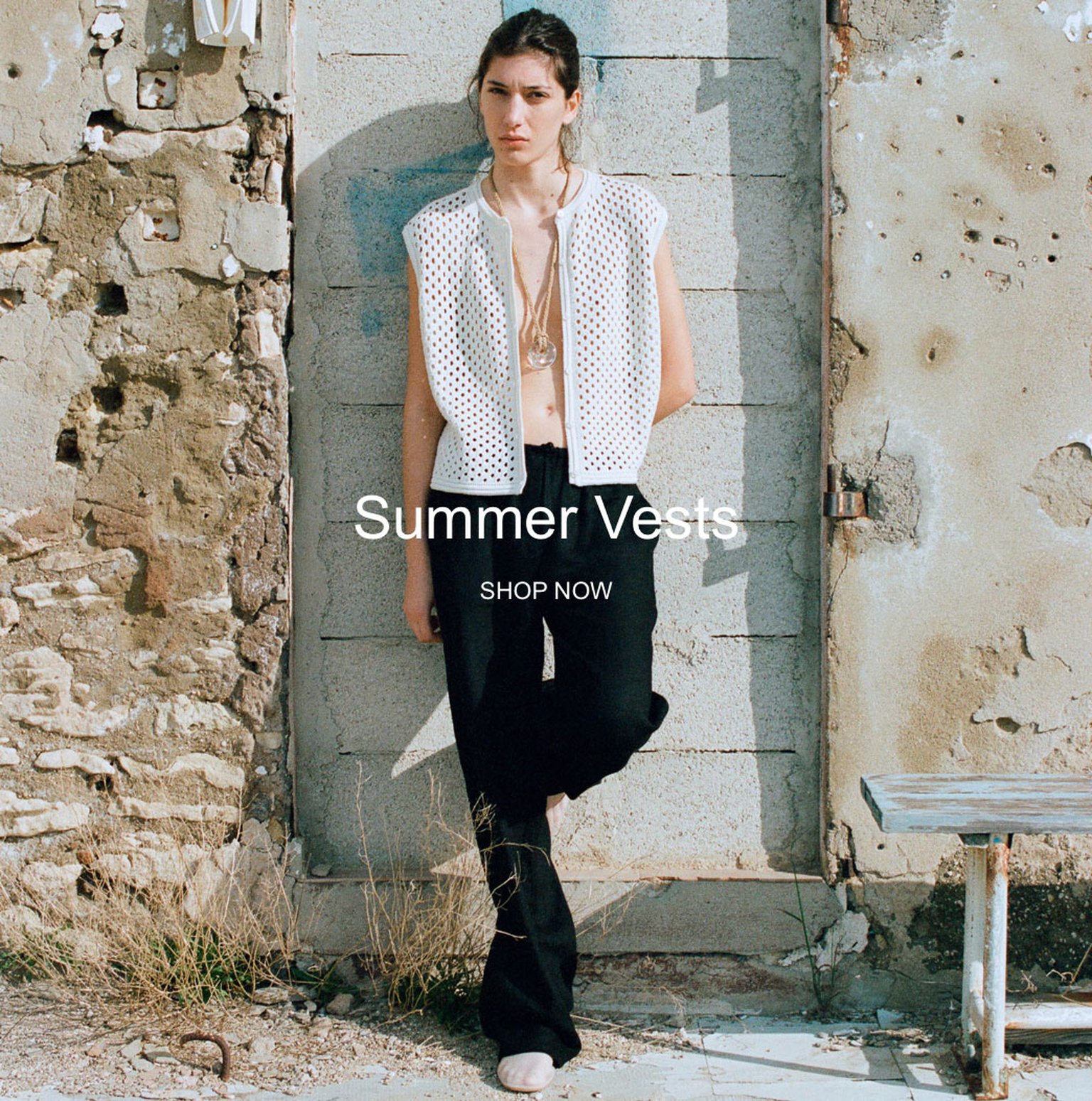 Person in a sleeveless white vest and black pants stands against a weathered wall. Text reads "Summer Vests, Shop Now."