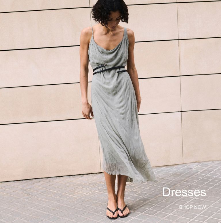 Woman in a flowing grey dress with a black belt stands on a tiled pavement against a beige wall. Text reads "Dresses, Shop Now."