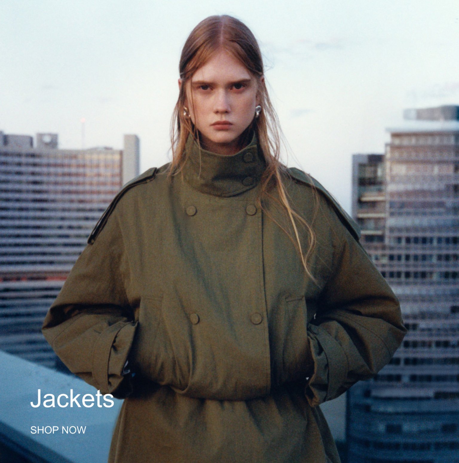 Person in a green double-breasted coat stands on a rooftop, with city buildings in the background. Text reads "Jackets SHOP NOW."