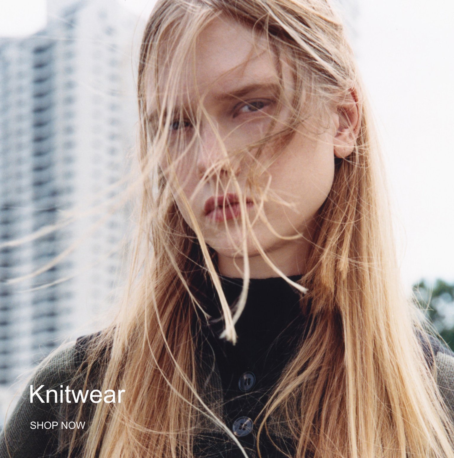Woman with long blonde hair blowing in the wind, wearing a dark top, stands outdoors with blurred buildings in the background. Text reads "Knitwear SHOP NOW".