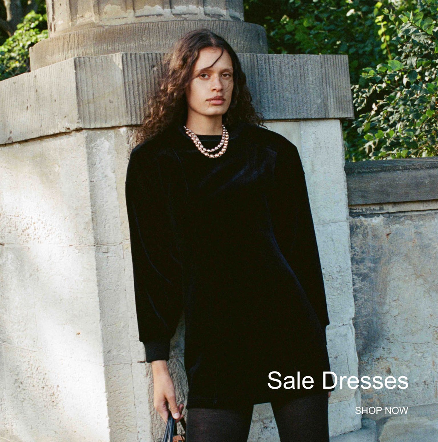 Person in black dress and pearl necklace stands by stone structure, surrounded by greenery. Text reads, "Sale Dresses, Shop Now."