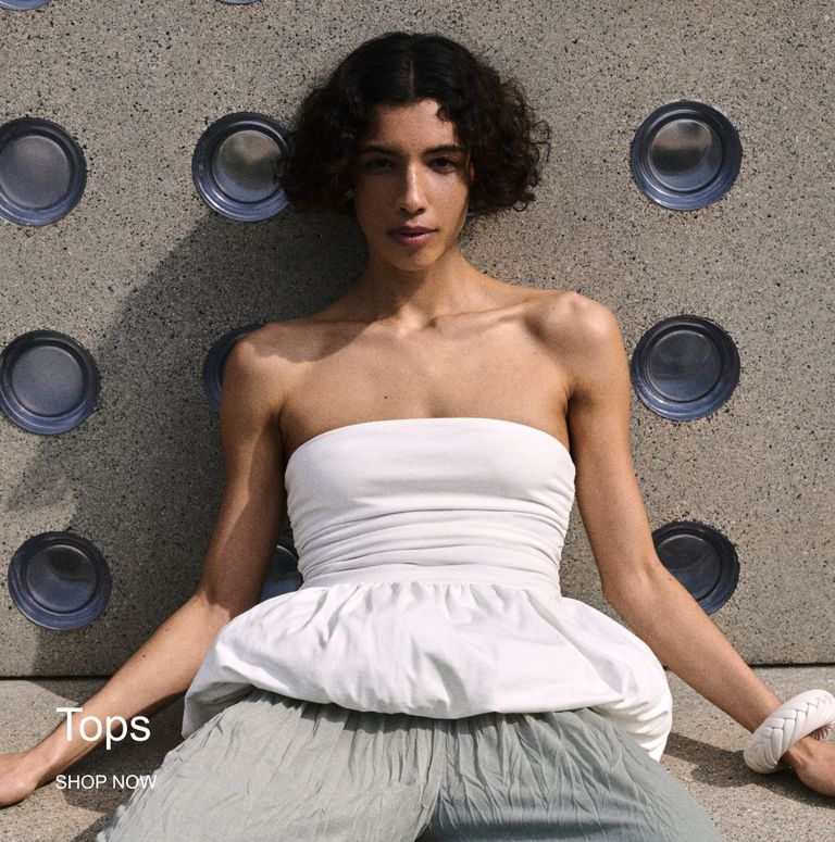 Person in a white strapless top and gray pants sits against a textured wall adorned with circular patterns. Text reads: "Tops SHOP NOW".
