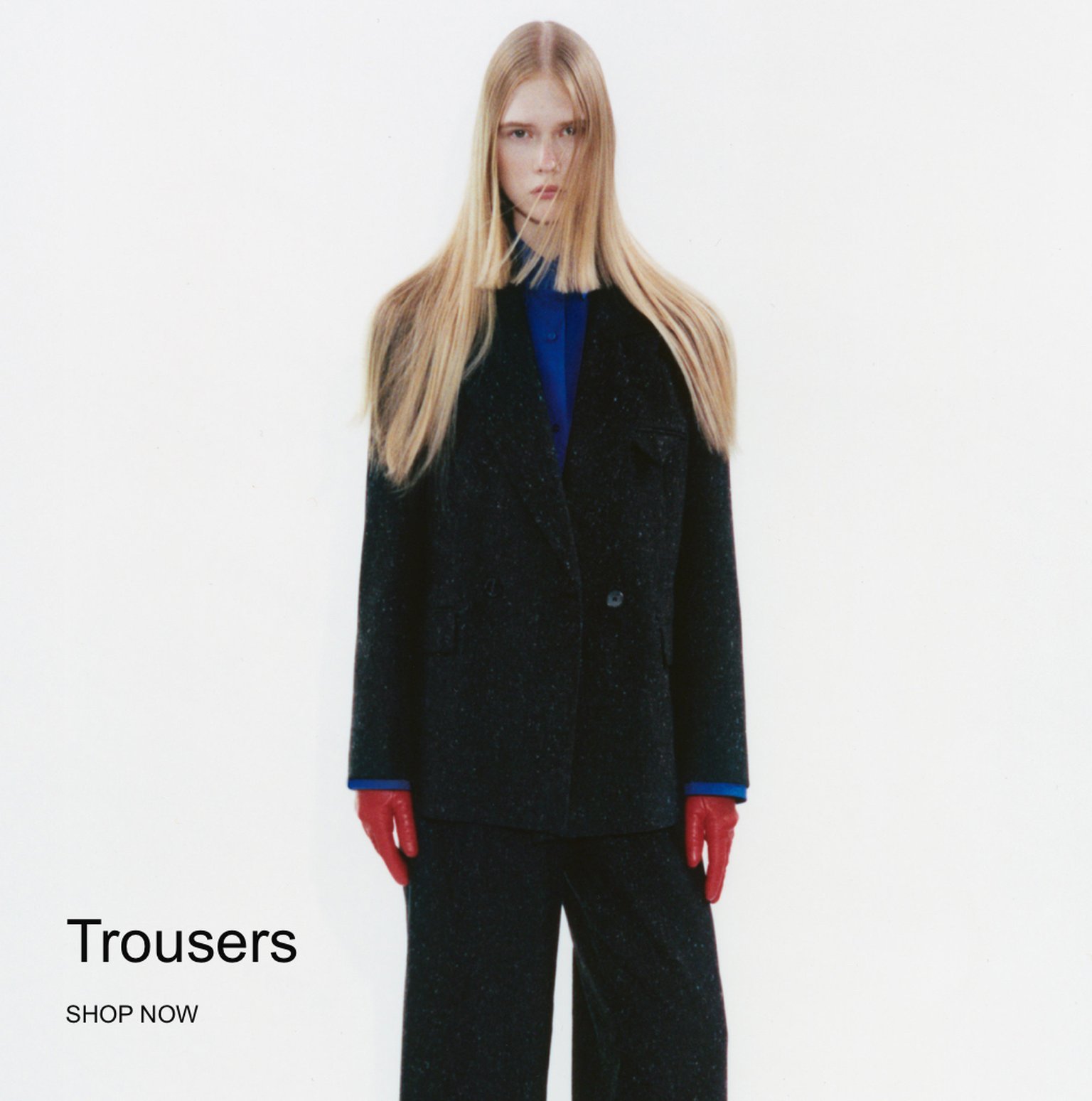 Person with long hair wearing a dark suit, blue shirt, and red gloves against a white background. Text reads "Trousers SHOP NOW".