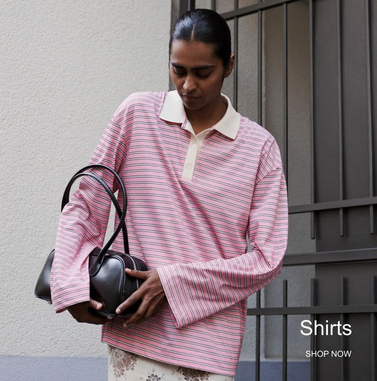 Person wearing a striped pink shirt and holding a black bag, standing near a gated wall. Text reads "Shirts SHOP NOW."