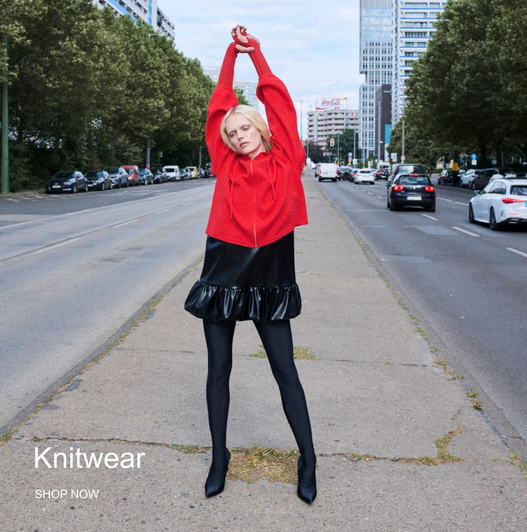Person in a red knit sweater and black skirt poses on an urban sidewalk with arms raised; text reads "Knitwear, Shop Now".