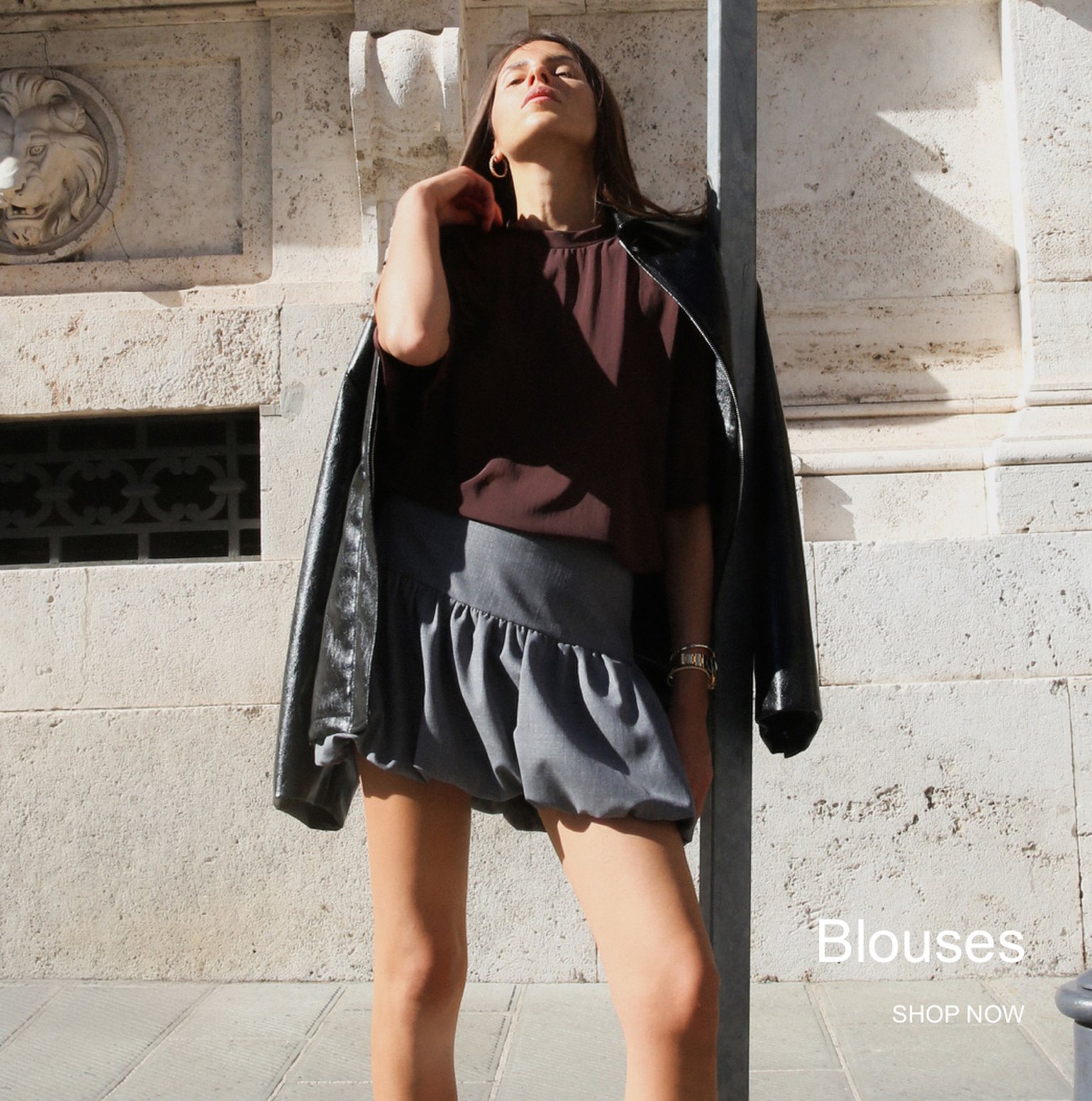 Woman posing confidently in sunlight, wearing a brown blouse, grey skirt, and black jacket draped over shoulders. Text reads "Blouses SHOP NOW."