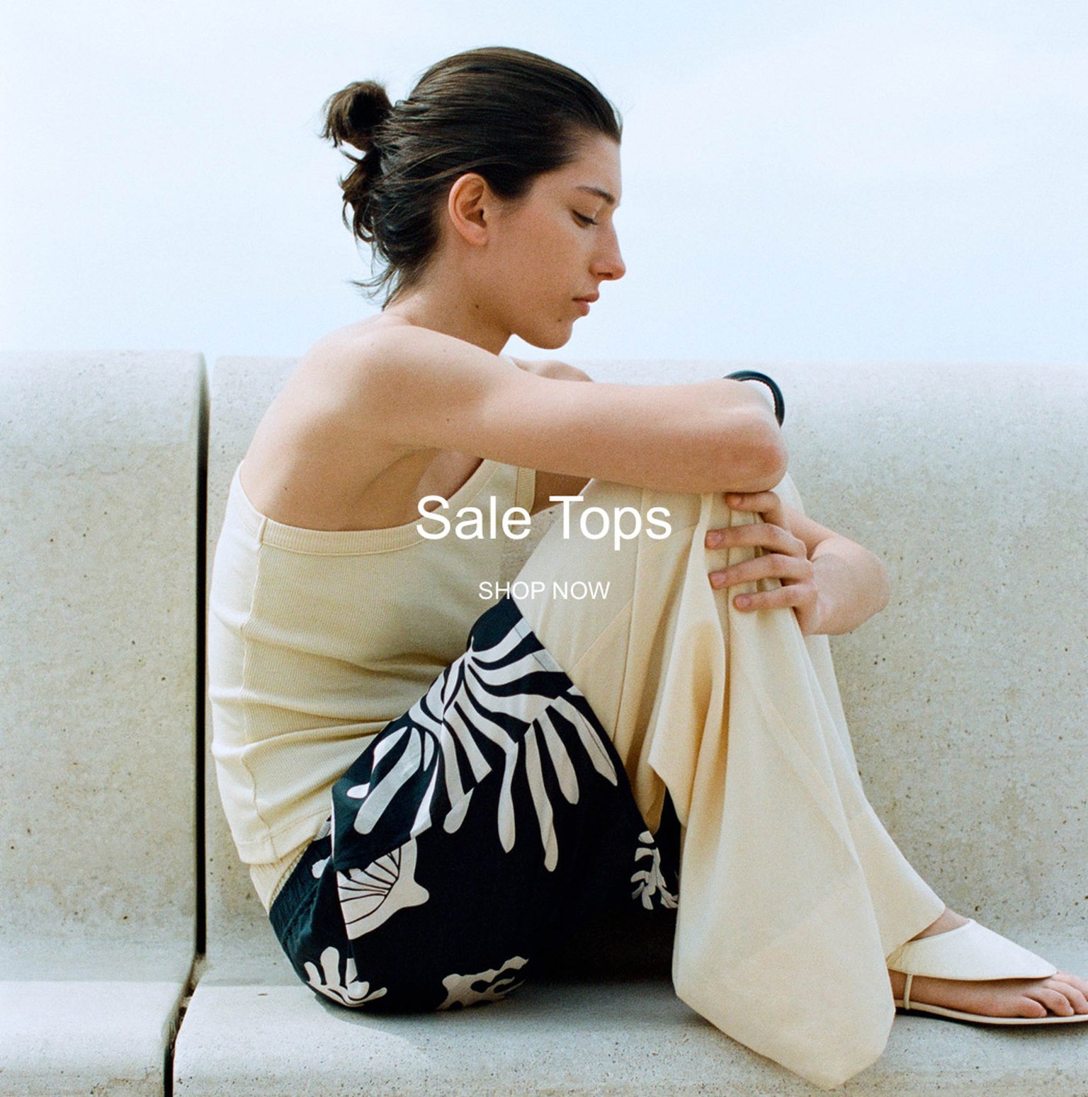 Woman sitting on a bench in a sleeveless top and patterned pants, with text overlay: "Sale Tops, Shop Now."
