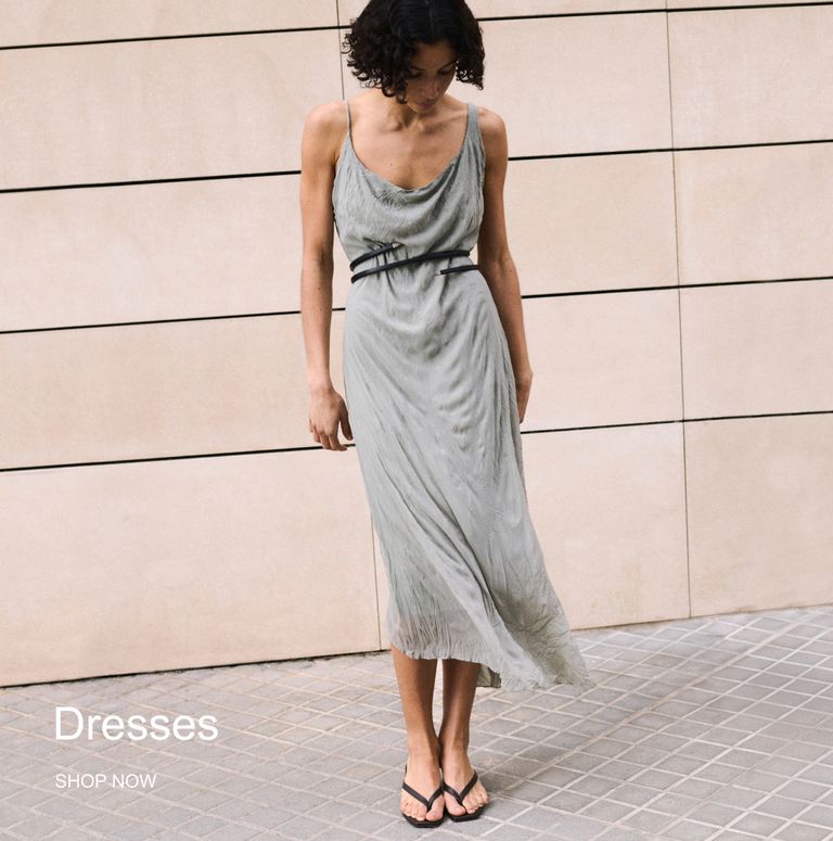 A woman in a light green slip dress, cinched with a thin belt, stands against a beige tiled wall. She wears black sandals and looks down.