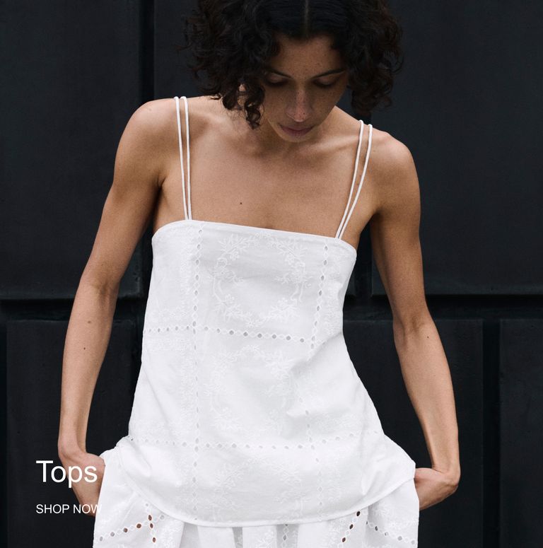 Woman in a white strappy top with eyelet detailing, standing against a dark background. Text reads "Tops, Shop Now."