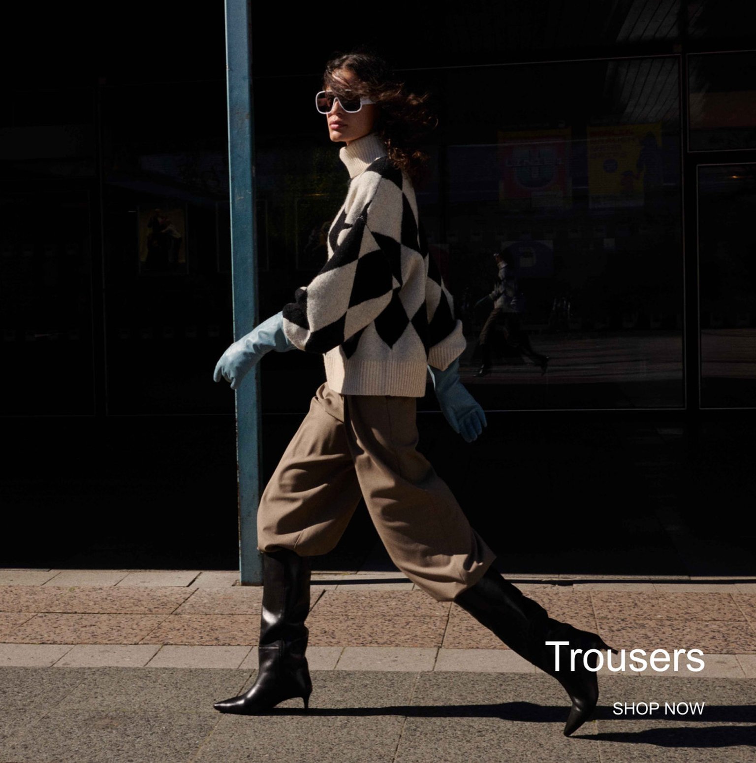 Fashionable person in checkered sweater, baggy trousers, and boots walking on a sunny day. Text reads "Trousers" and "Shop Now."