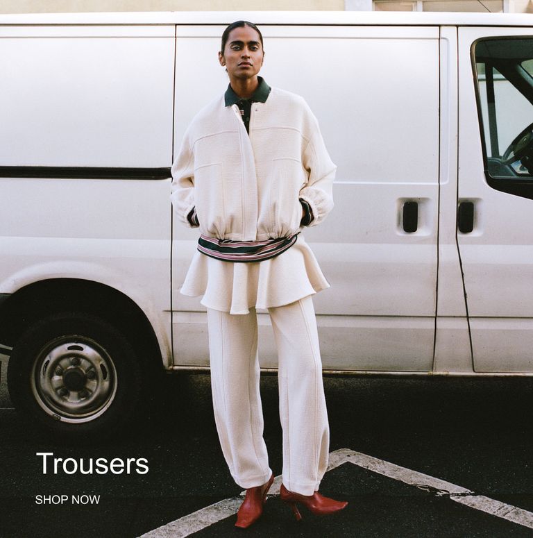 Person in a white outfit with red shoes stands in front of a white van. Text reads "Trousers SHOP NOW."