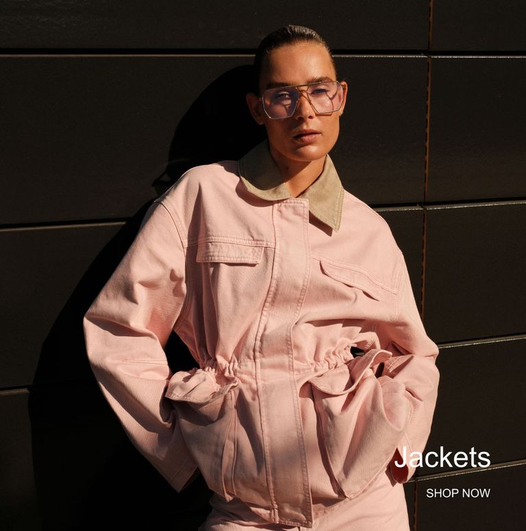 Person wearing a light pink jacket with large pockets, standing against a dark wall. Text reads "Jackets, Shop Now."