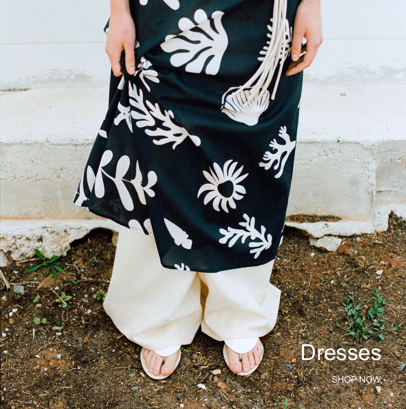 Person wearing a black dress with white floral patterns over cream pants, standing on soil. Text reads "Dresses SHOP NOW."