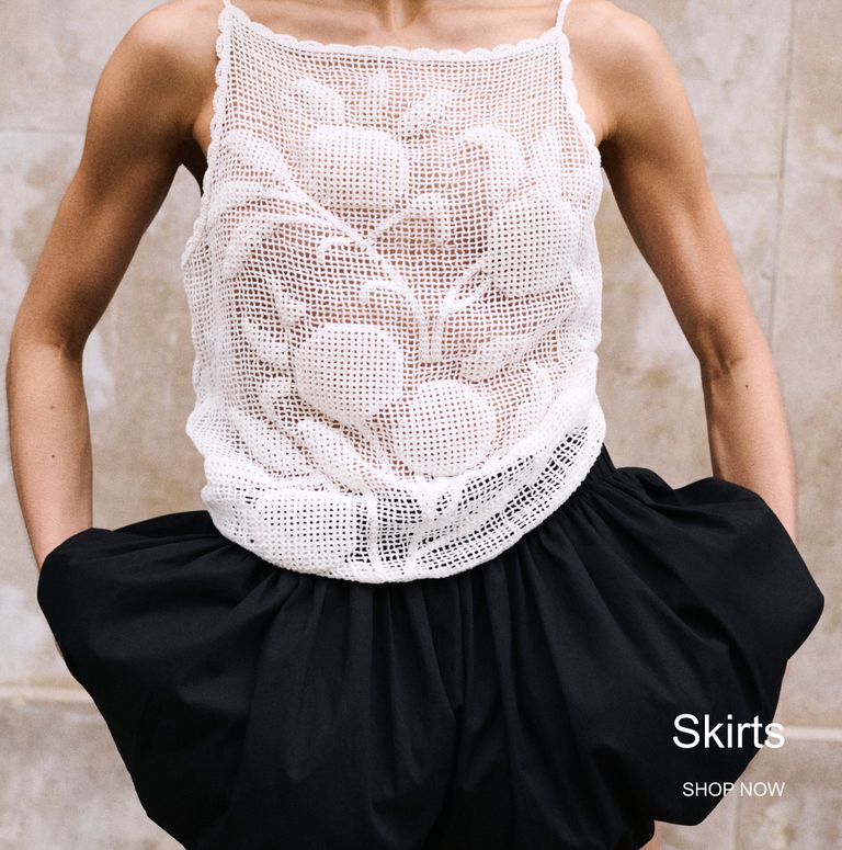 A person wearing a white crochet tank top and a black skirt, standing with hands in pockets. The word "Skirts" and "SHOP NOW" are visible.