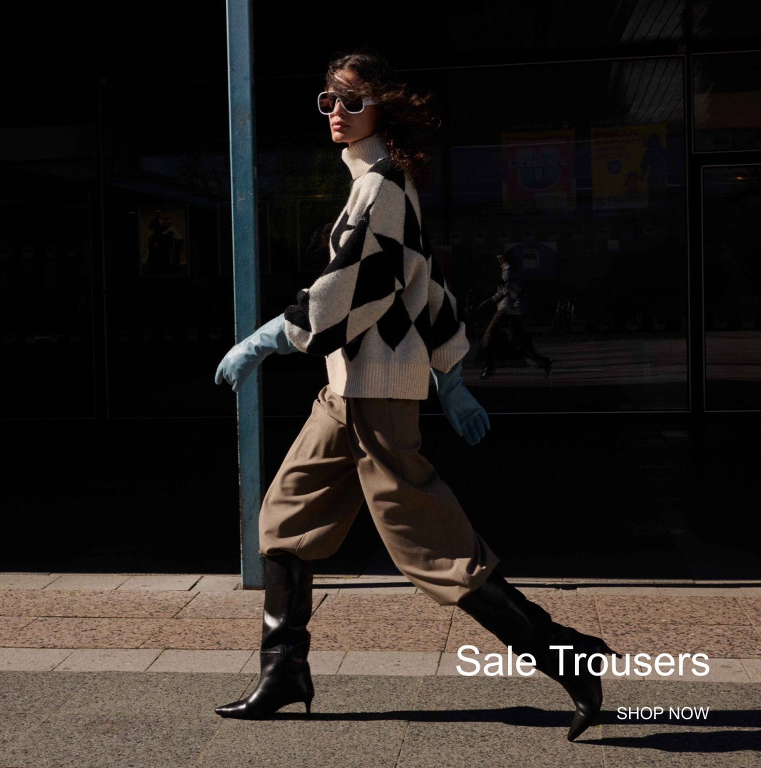 Fashionable person in sunglasses, black-and-white sweater, wide trousers, boots, and blue gloves, walking by a glass storefront. Text: "Sale Trousers, SHOP NOW".