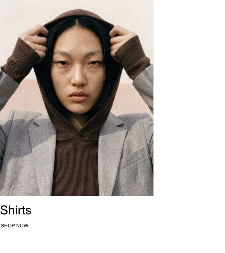 A person wearing a brown hoodie under a gray blazer, holding the hood with both hands, with text "Shirts" and "Shop Now" at the bottom.