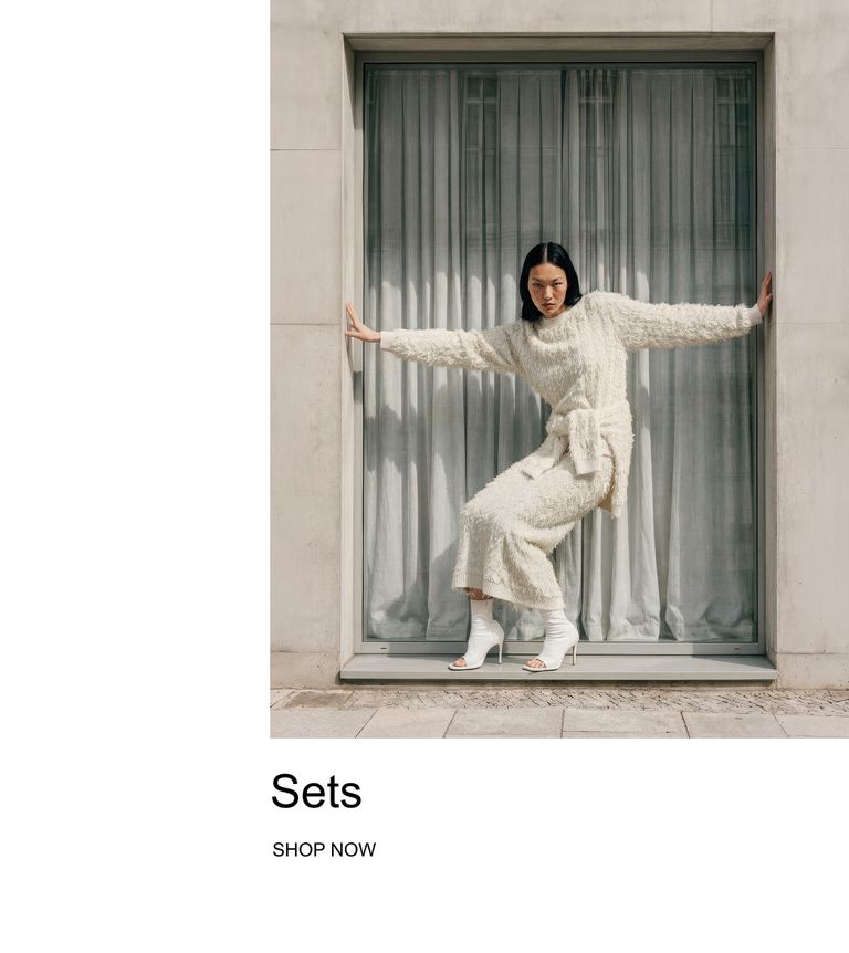 A person in a fluffy white knit set poses in a window, wearing heeled sandals. Text below reads "Sets, Shop Now."