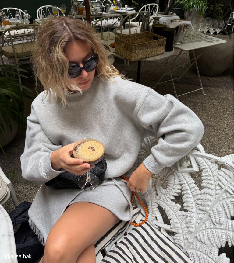 Person in a gray outfit and sunglasses holds a cocktail in a garden café, sitting on a white ornate chair with striped cushions.
