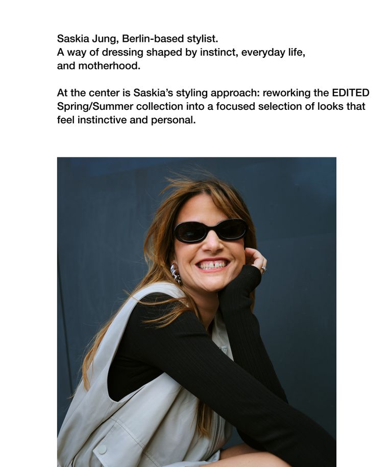 Woman in sunglasses smiling, wearing a black top and light vest, sitting against a dark blue background, looking relaxed and stylish.