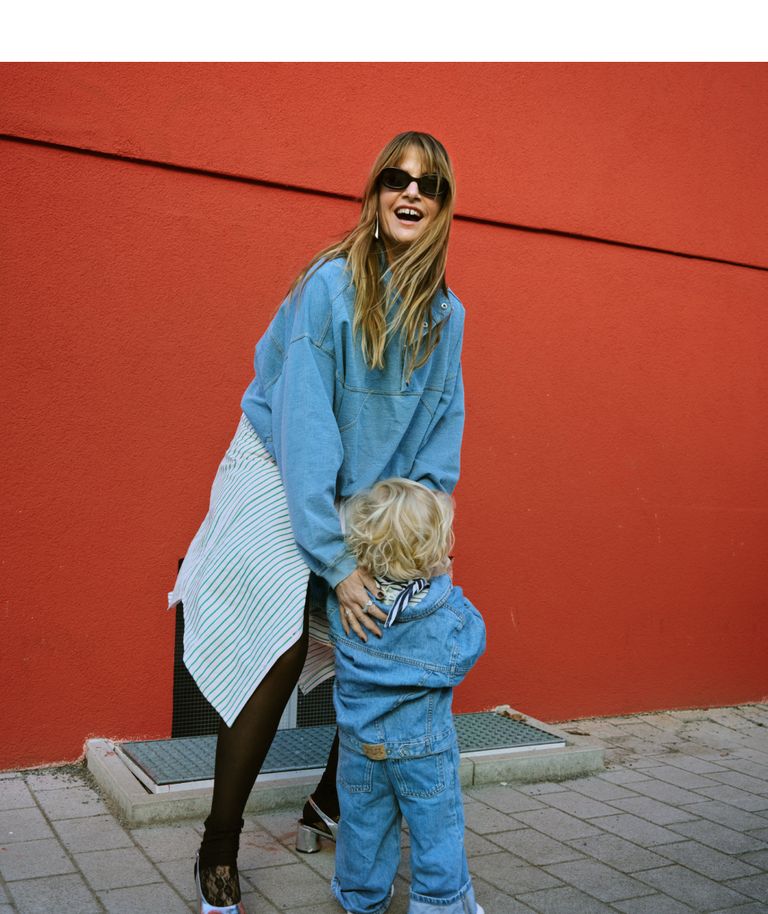 Woman in sunglasses and blue jacket joyfully embraces a child in denim against a red wall.