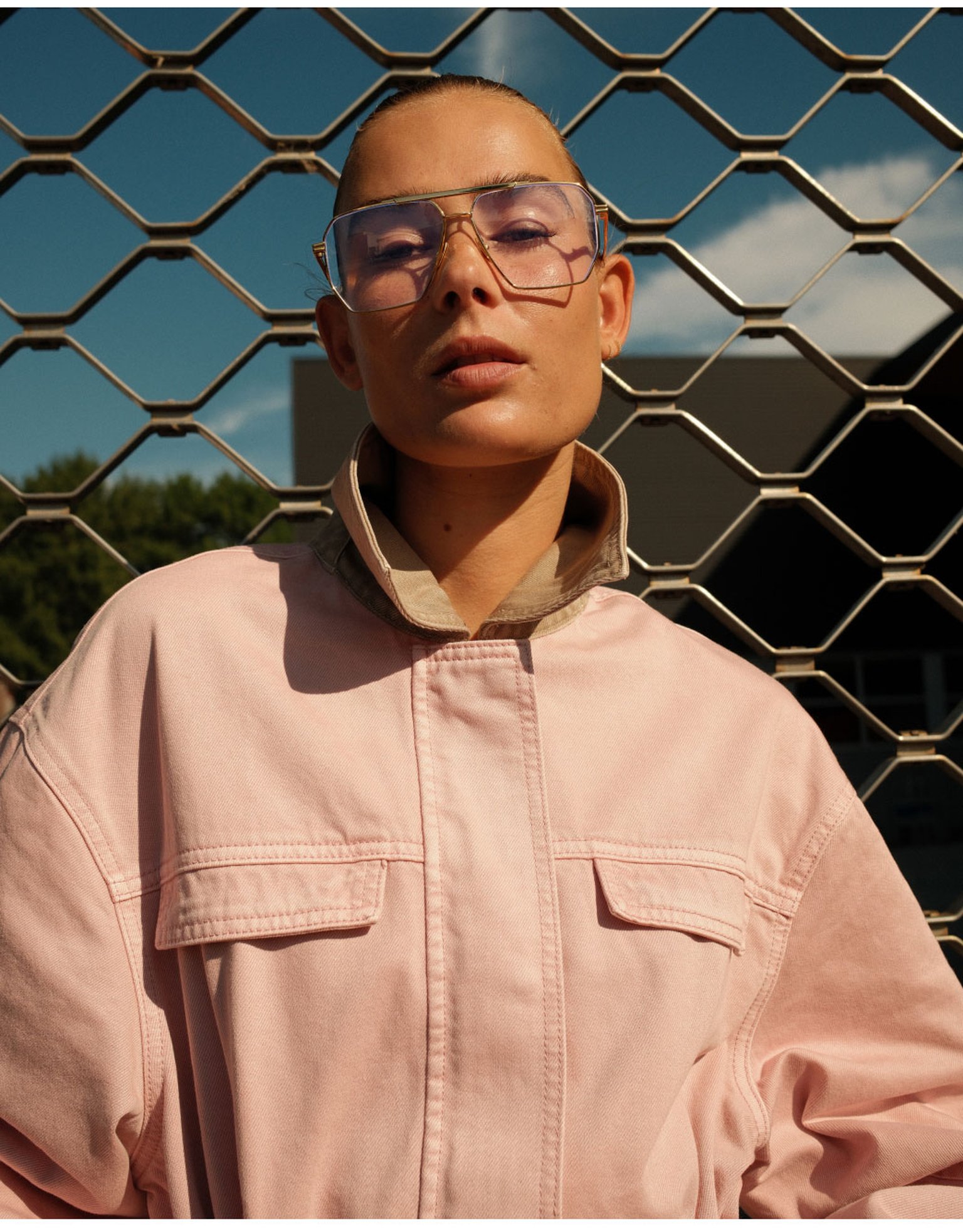 Person wearing a pink jacket and tinted glasses, standing in front of a chain-link fence under a clear sky.