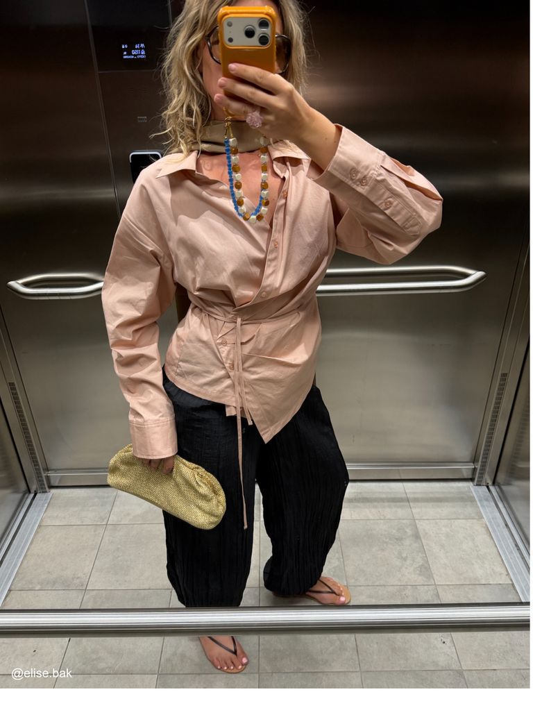 Person in an elevator taking a selfie, wearing a pink wrap shirt, black wide-leg pants, and sandals, holding a gold clutch and phone.