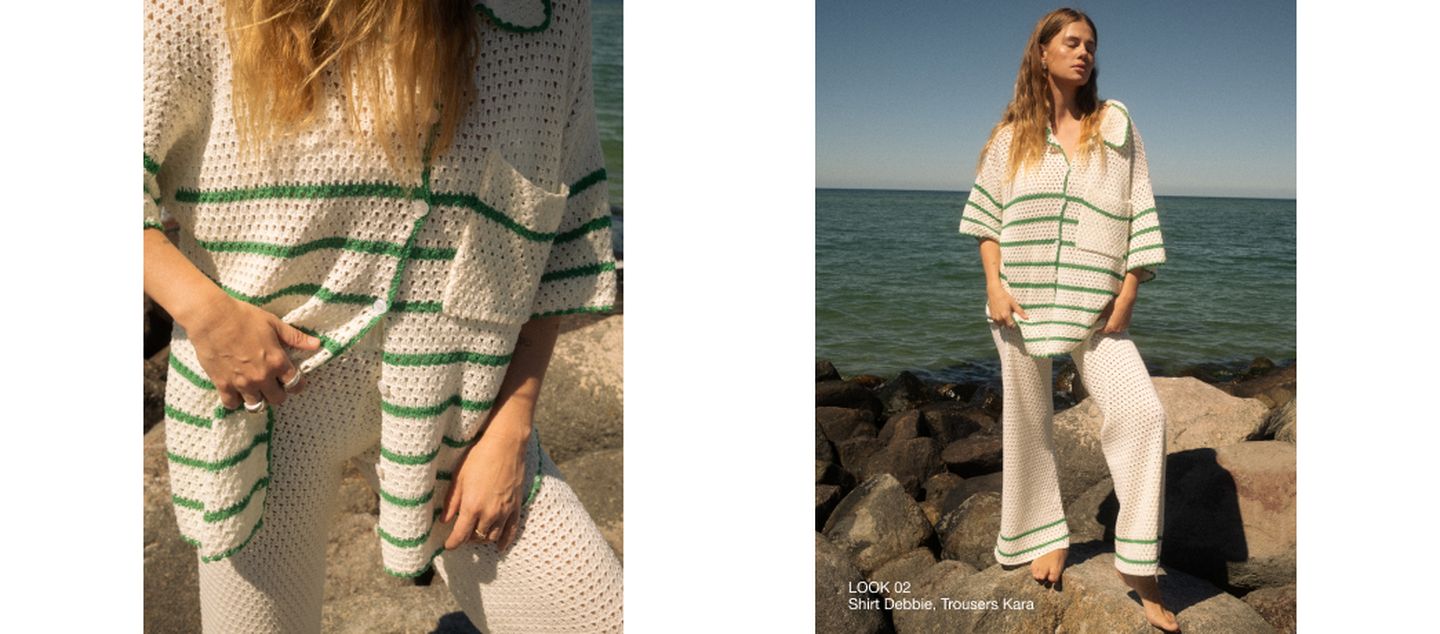 Woman in a loose-knit white crochet outfit with green stripes, posing by rocks and the sea under a clear blue sky.