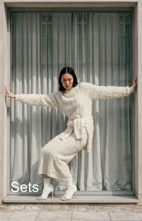 A person in a white textured knit outfit poses with arms outstretched in a window frame, wearing white high-heeled shoes. Text reads "Sets" and "Shop Now."