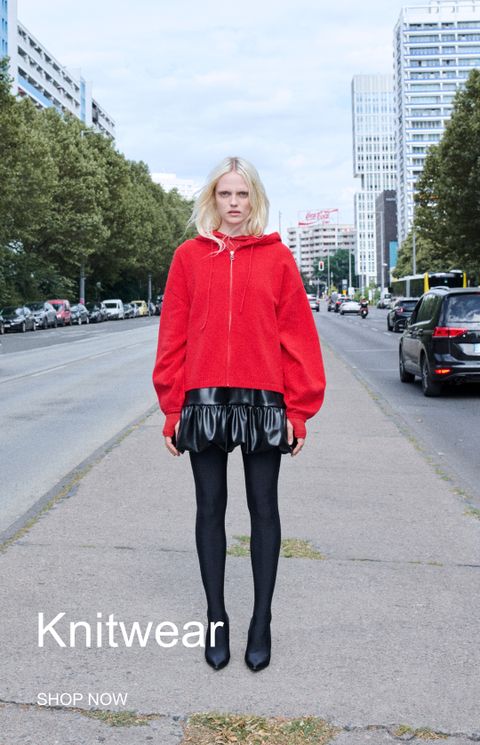 A person stands on a city street wearing a red zip-up sweater, black skirt, and tights. Text reads "Knitwear. Shop Now."
