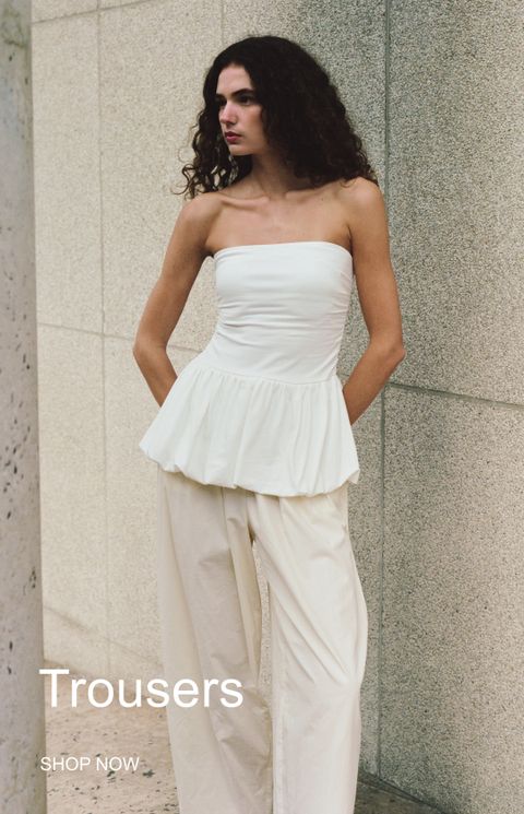 Woman in a white strapless top and cream trousers leans against a stone wall. Text reads "Trousers" and "SHOP NOW" in the corner.