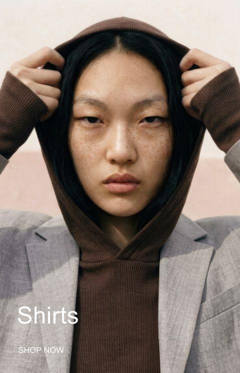 Person with freckles pulls hoodie over head, wearing a gray blazer. Text reads "Shirts, Shop Now."