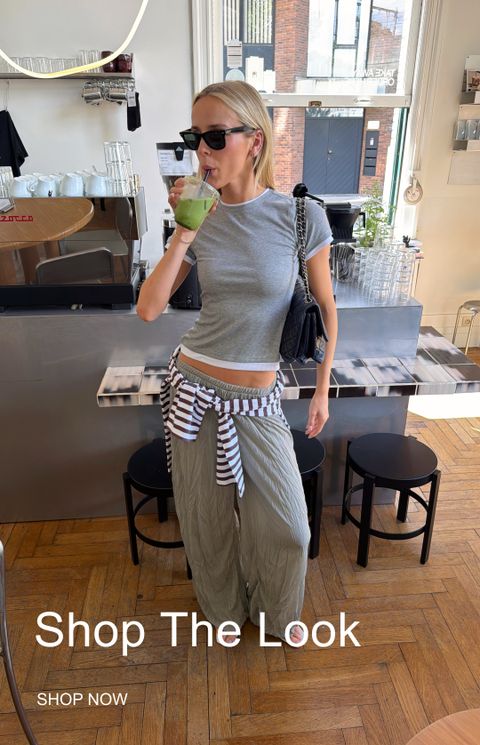Woman in a cafe drinking a green smoothie, wearing sunglasses, a grey T-shirt, olive pants, and a striped shirt tied at the waist. "Shop The Look" text.
