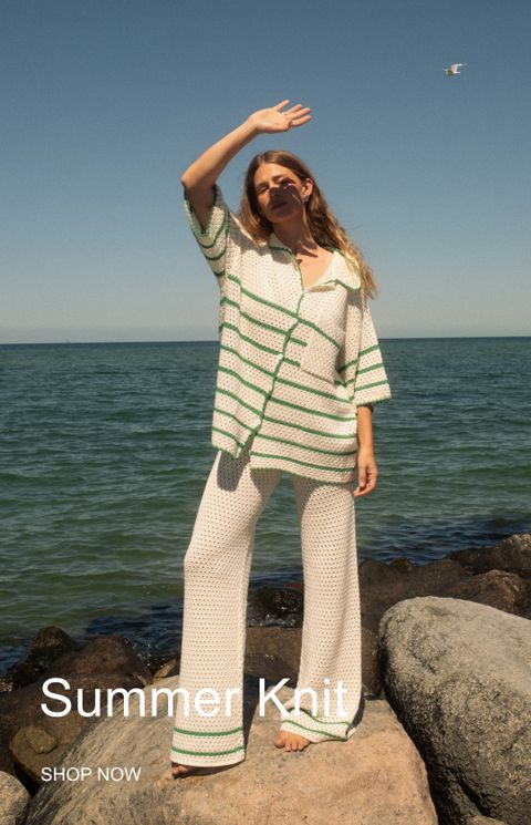 A woman in a light summer knit outfit stands on rocky shoreline, shielding her eyes from the sun, with the ocean in the background.