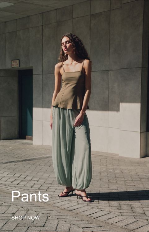 A person in a brown top and loose green pants stands in front of a modern building. Text overlay reads "Pants" and "Shop Now."