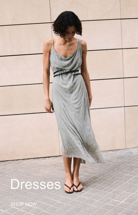 Woman in a flowing grey dress with a black belt stands on a tiled pavement against a beige wall. Text reads "Dresses, Shop Now."