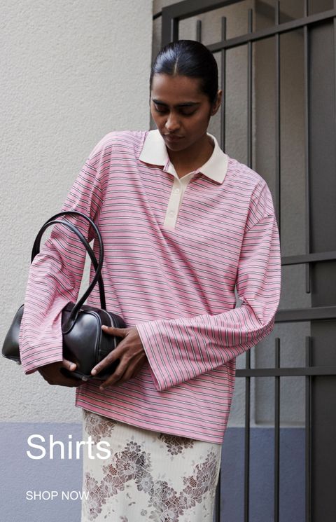 Person wearing a striped pink shirt and holding a black bag, standing near a gated wall. Text reads "Shirts SHOP NOW."